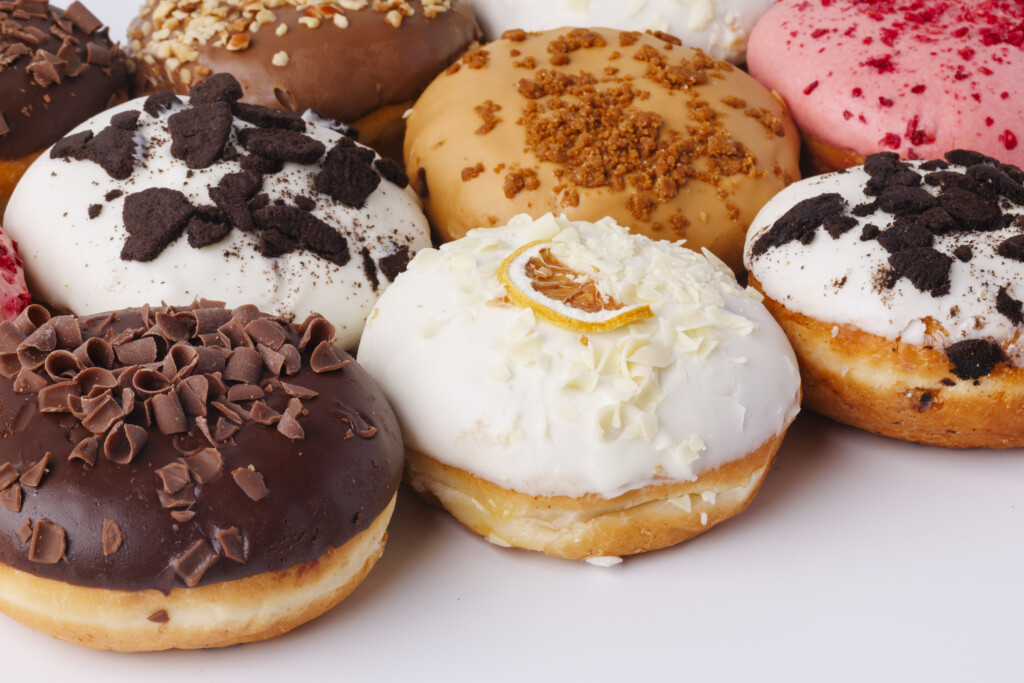Close-up assortment of colorful Pie Face donuts, featuring various toppings like chocolate shavings, crushed nuts, white icing, pink icing, and cookie pieces, representing the sweet treats range.